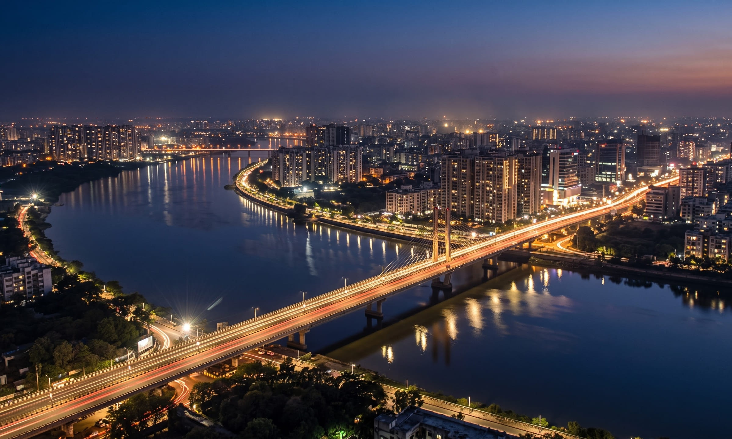 The Surat skyline and the Tapi river at blue hour, with traffic light trails on a bridge and calm water reflections.